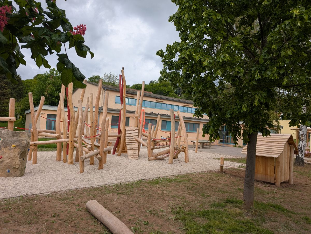 Naturnaher Spielplatz mit Holzkletterelementen und Spielhaus auf einem Schulhof